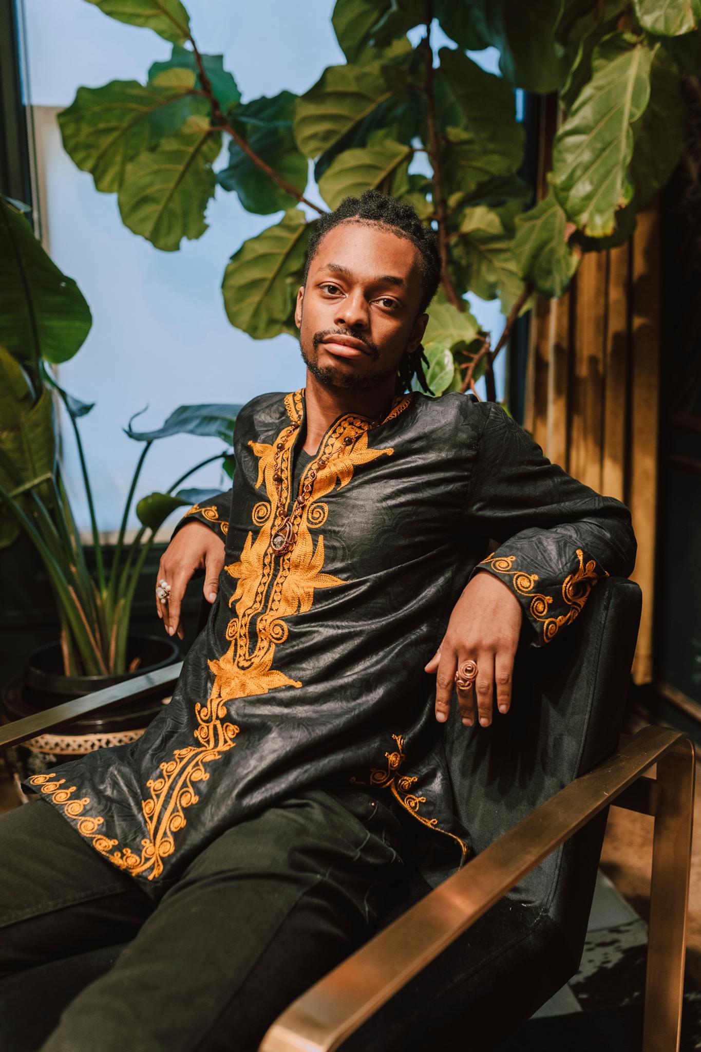 A fashionable man in a dashiki sits indoors surrounded by lush plants.