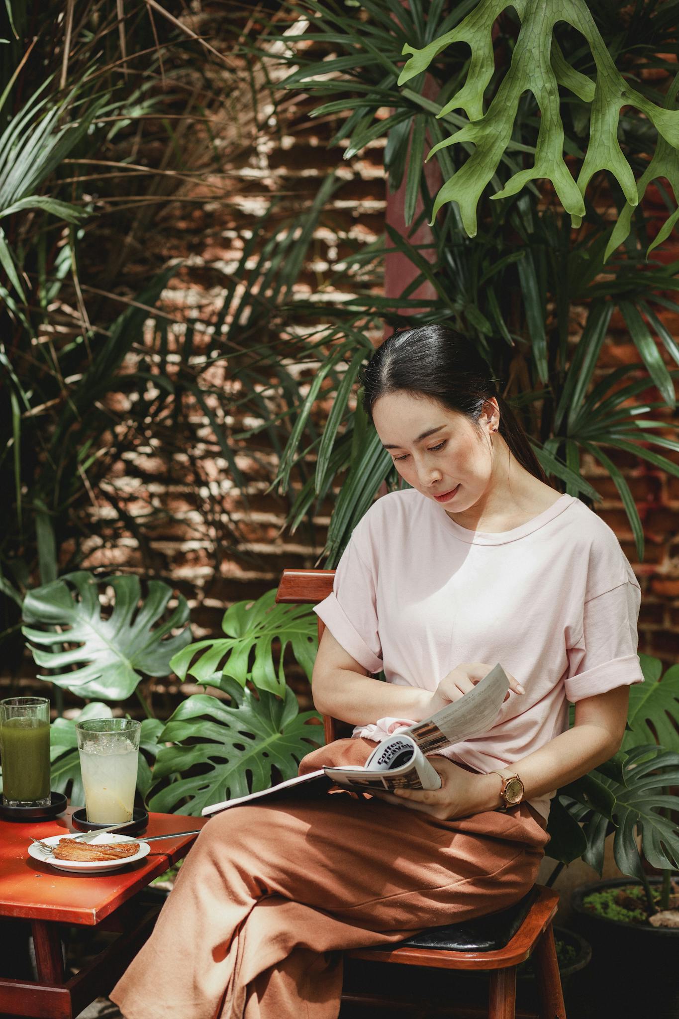 Calm Asian woman in casual clothes sitting in cafe and reading interesting magazine against green plants on brick wall